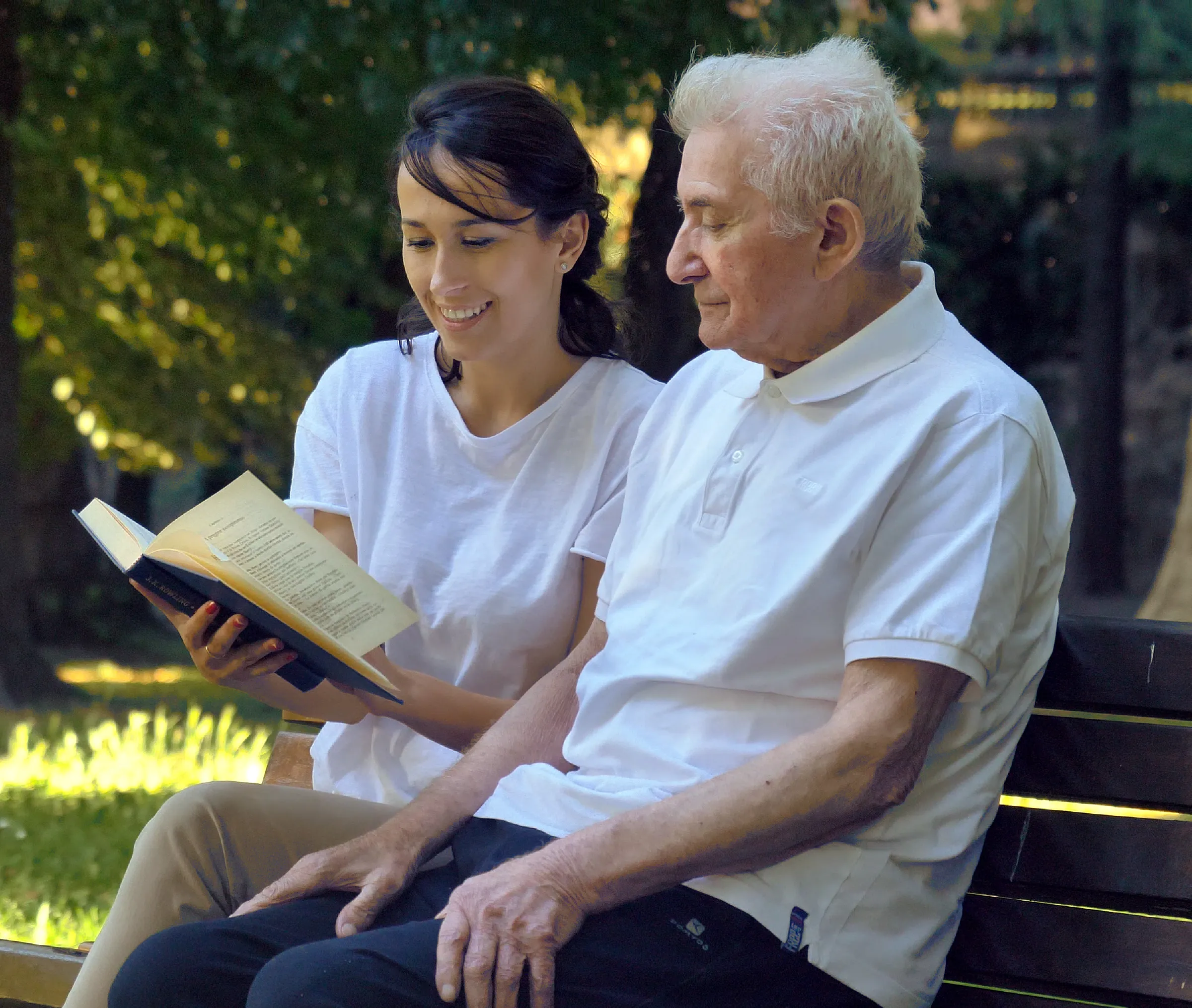 man on bench outside of memory care community with woman reading to him